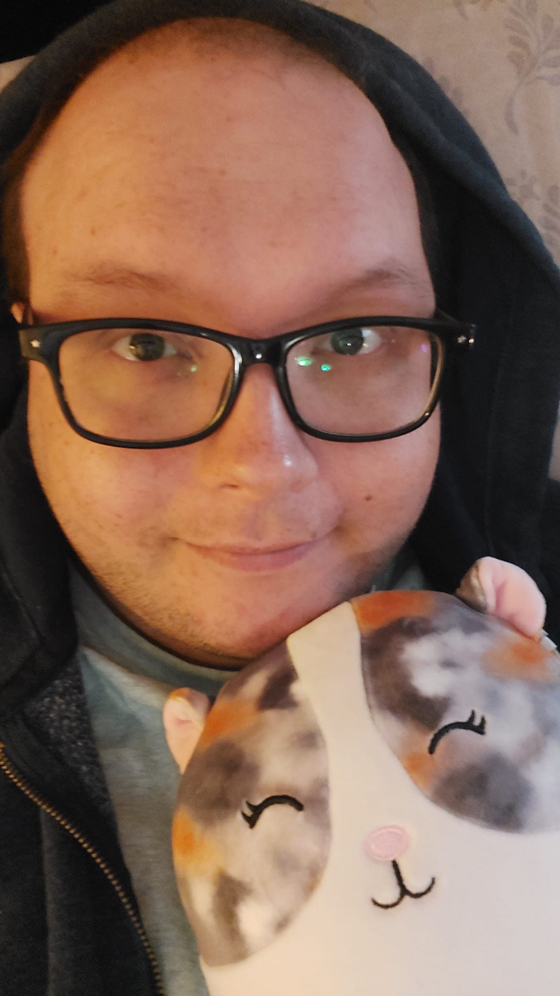 A man lies in bed, fully dressed, holding a small hamster Squishmallow, both look happy